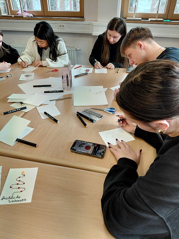 Eine Gruppe junger Menschen sitzt um einen Tisch und zeichnet Weihnachtsmotive auf Karten, Stifte und Papier liegen verstreut auf der Tischfläche.