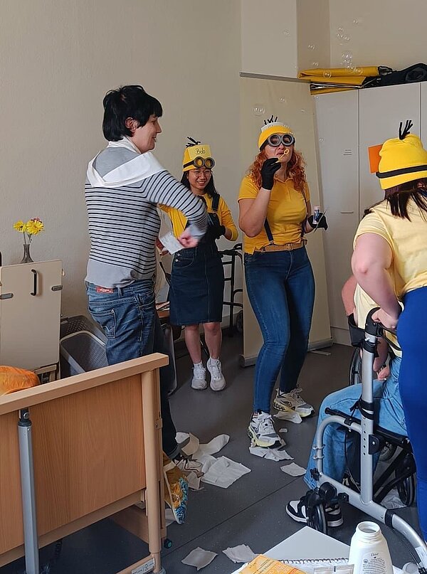 Operation Krankmacher: Die Mission der Minions Schülerinnen und Schüler der Pflege im Minionkostüm haben viel Spaß während der Darstellung der Geschichte. Sie blasen Seifenblasen und wickeln sich mit Verbandsmaterial ein. Eine Person sitzt im Rollstuhl während eine andere sie schiebt.