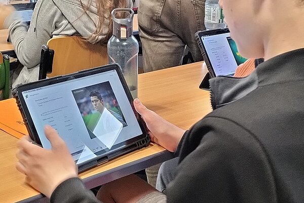 In einem Klassenraum der Euro Akademie Görlitz sitzt ein Schüler an einem Holztisch und hält ein Tablet in den Händen. Auf dem Bildschirm ist eine Website mit einem Bild und Text zu sehen, die sich thematisch mit Fake News auseinandersetzt. Im Hintergrund sind weitere Personen an Tischen erkennbar, die ebenfalls Tablets nutzen. Auf dem Tisch steht eine durchsichtige Trinkflasche mit dunklem Deckel. Die Szene vermittelt den Einsatz digitaler Medien im Unterricht und verdeutlicht das medienpädagogische Projekt zur Förderung von Medienkompetenz im Umgang mit Falschinformationen.
