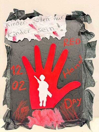 Ein dunkles Plakat mit einem großen roten Handabdruck in der Mitte. Darauf steht ‚12.02‘ und ‚RED Hand Day‘. Im Handabdruck ist die Silhouette eines bewaffneten Soldaten zu sehen. Oben auf dem Plakat steht ‚Kinder sollen nur Kinder sein‘.