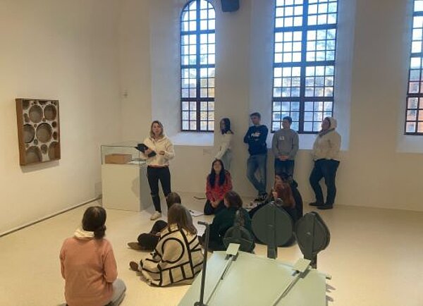 Eine Gruppe von Personen nimmt an einer Führung in einem Kunstmuseum teil. Einige sitzen auf dem Boden, während andere stehen und einer Person zuhören, die vorne spricht. Im Hintergrund sind Kunstwerke an den Wänden zu sehen.