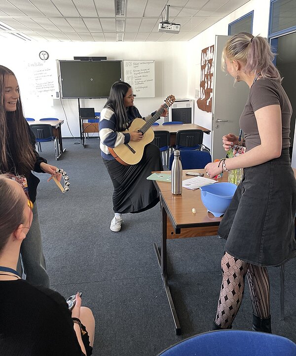 Erfolgreicher Tag der offenen Tür Auszubildende und Besucherinnen und Besucher in einem Klassenraum stehen zusammen, eine Auszubildende hat eine Akustikgitarre in der Hand und spielt etwas vor.