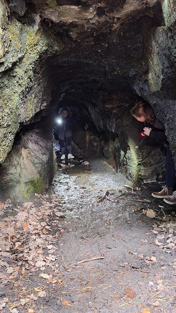 Zwei Personen mit Taschenlampen erkunden eine dunkle, feuchte Höhle mit felsigen Wänden, während der Boden mit Blättern und Wasser bedeckt ist.