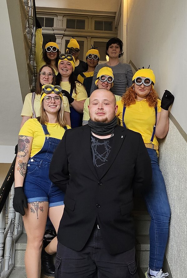 Die letzte Schulwoche der PFM 22: Ein unvergesslicher Abschluss Eine Gruppe junger Erwachsener steht verkleidet als Minions auf einer Treppe. Sie tragen gelbe Mützen, Brillen, gelbe T-Shirts und blaue Latzhosen. Vor der Gruppe steht ein Mann in schwarzem Anzug mit ernster Miene.