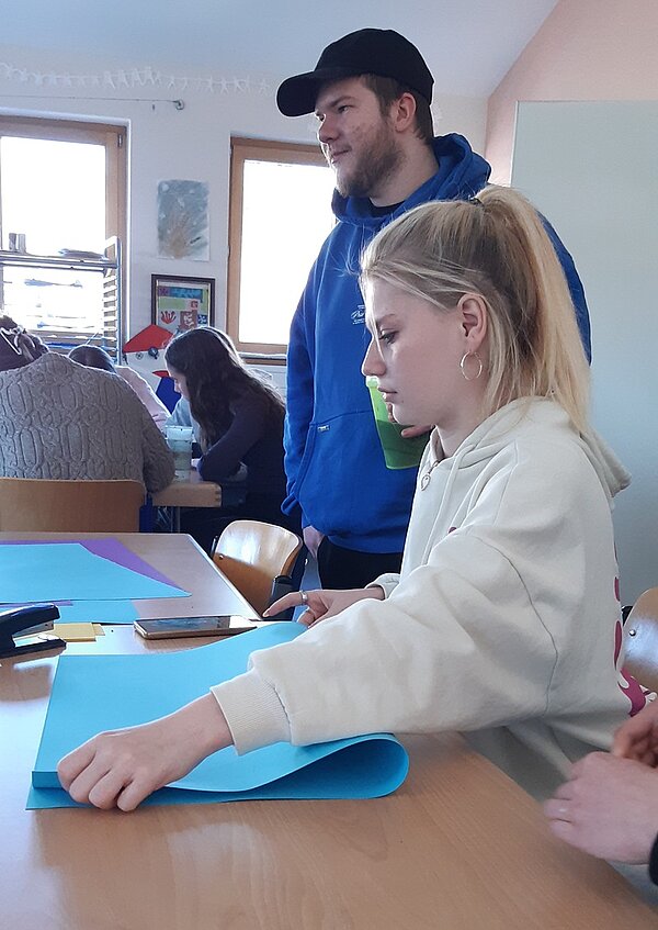 Einstimmung auf Ostern Eine weibliche Auszubildende mit Pferdeschwanz faltet ein großes Blatt blaues Tonpapier in der Mitte, neben ihr steht ein männlicher Mitazubi in blauem Kapuzenpullover und mit schwarzer Badecap auf dem Kopf.