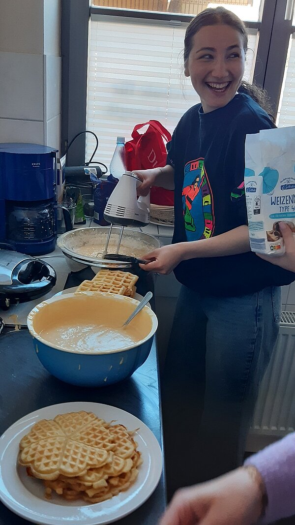 Einstimmung auf Ostern Eine weibliche Auszubildende steht in der Schulküche, vor ihr eine volle Schüssel mit Waffelteig, und rührt den Teig mit dem Mixer lächelnd und gut gelaunt. Auf dem Tisch liegen bereits fertige Waffeln auf einem Teller gestapelt.