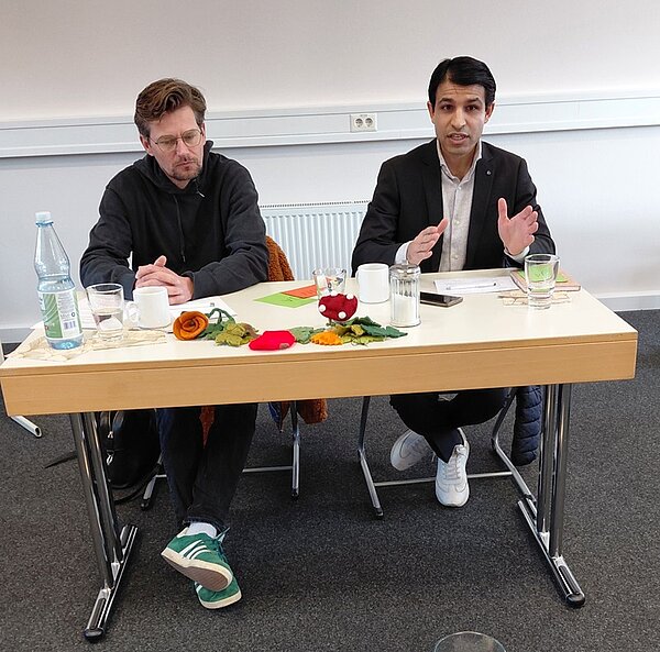 Die beiden männlichen Politiker sitzen nebeneinander an einem rechteckigen Tisch in einem Seminarraum der Euro Akademie Tauberbischofsheim. Auf dem Tisch stehen eine Glasflasche mit Wasser, mehrere Gläser, eine Kaffeetasse, ein Notizblock, ein Kugelschreiber sowie herbstliche Dekoration mit künstlichen Blättern und einer Stoffrose. Im Hintergrund verlaufen eine weiße Wand mit Steckdose und ein Heizkörper. Beide Personen tragen legere Kleidung, eine Person einen dunklen Anzug mit weißem Hemd, die andere eine dunkle Jacke und grüne Turnschuhe. Die Szene vermittelt den Eindruck eines Gesprächs oder einer Diskussionsrunde im Rahmen eines offiziellen Besuchs zweier Politiker an der Bildungseinrichtung.