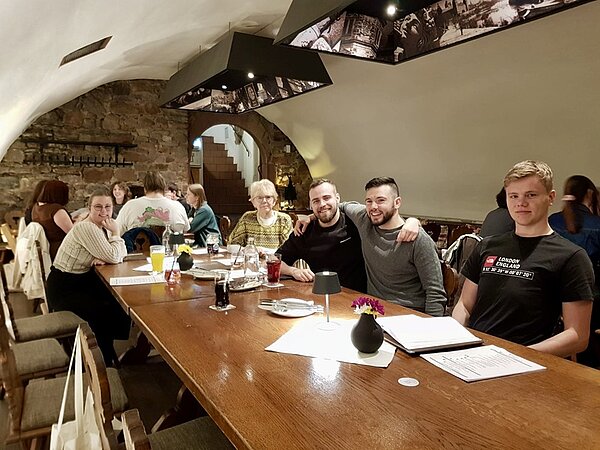 Alumnitreffen 2025 Drei männliche Auszubildende und eine weibliche ehemalige Auszubildende der Euro Akademie Aschaffenburg sitzen mit einer Lehrerin an einem Tisch im Restaurant und posieren lächelnd für ein Gruppenfoto. Vor ihnen liegen Speisekarten, Gläser mit verschiedenen Getränken, Besteck und eine kleine Tischlampe.