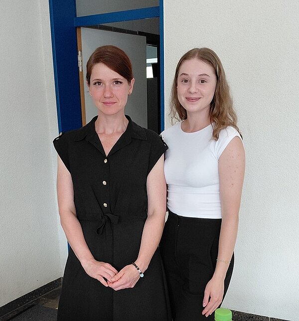 Zwei Absolventinnen posieren für ein Foto. Eine trägt das braune Haar zum Zopf und trägt ein schwarzes Kleid, die andere trägt die blonden Haare offen, ein weißes Top und ein schwarze Hose.