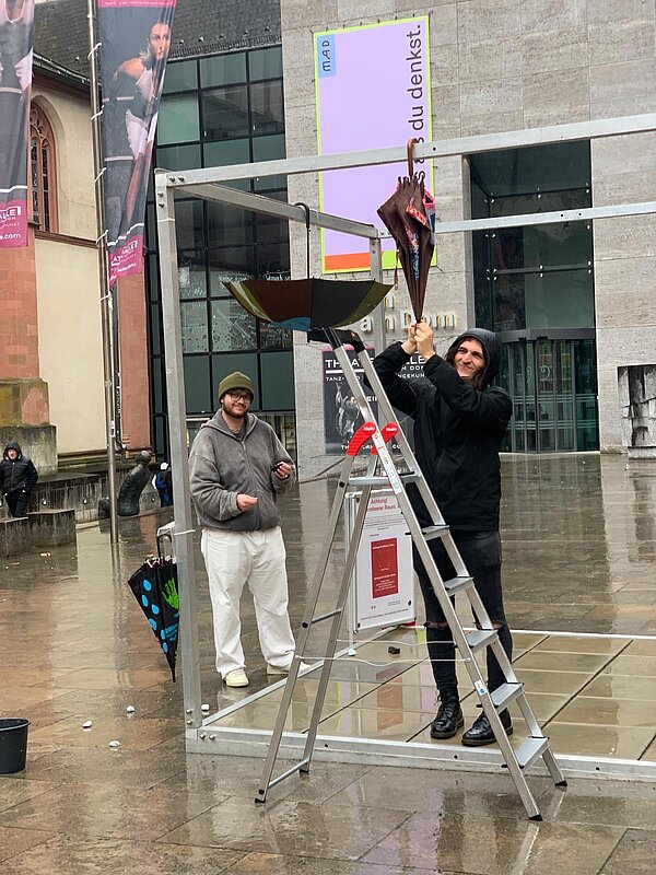 Auf einem verregneten Platz hängen zwei Menschen mit einer Leiter Regenschirme an einen Metallrahmen.