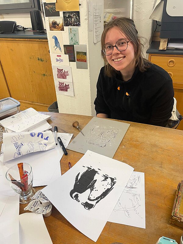 Eine Person mit Brille und mit einem schwarzen Hoodie bekleidet sitzt lächelnd an einem Holztisch mit Zeichenmaterialien und Skizzen, darunter ein Druck in schwarz-weiß von zwei Pinguinen. An der Wand im Hintergrund hängen aufgehängte Arbeiten und Notizen.