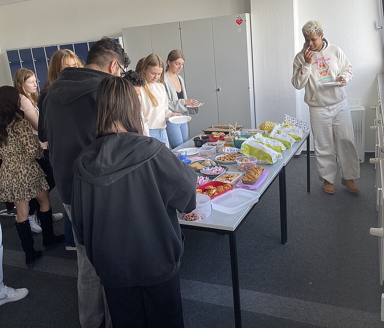 Diversity-Tag: internationaler Brunch an der Euro Akademie Aschaffenburg Ein Tisch voller leckerer Snacks, wie Salate, Knabberplatten, Rohkost und orientalische Köstlichkeiten. Herum stehen Schülerinnen und Schüler der Euro Akademie Aschaffenburg.