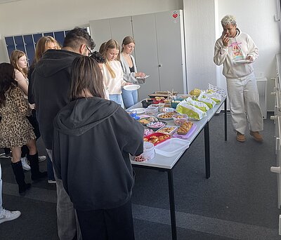 Ein Tisch voller leckerer Snacks, wie Salate, Knabberplatten, Rohkost und orientalische Köstlichkeiten. Herum stehen Schülerinnen und Schüler der Euro Akademie Aschaffenburg.