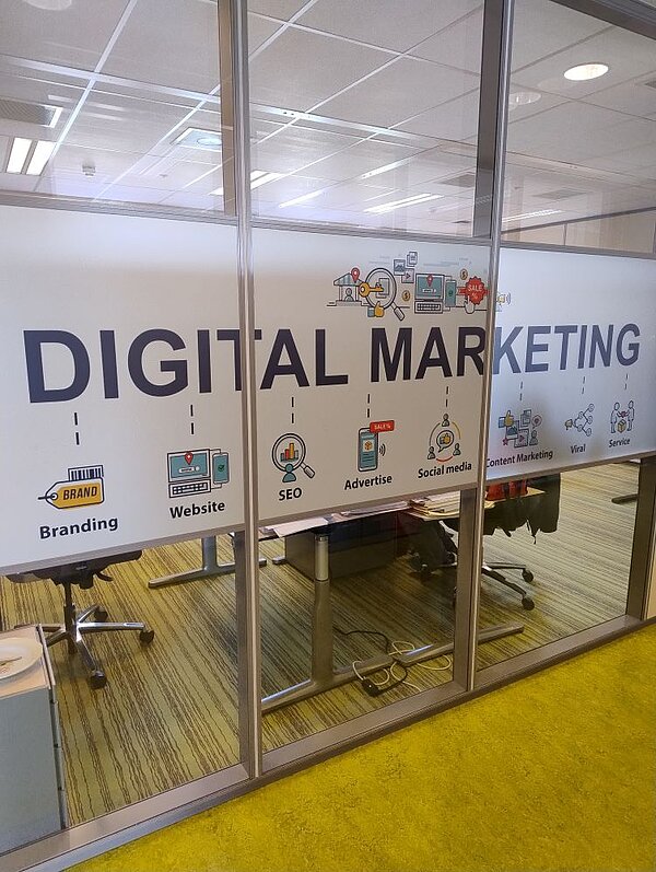 Eine Glastür mit der Aufschrift ‚DIGITAL MARKETING‘ und verschiedenen Symbolen für Social Media, SEO, Branding und Content Marketing. Durch die Tür sieht man in einen modernen Büro- oder Seminarraum.