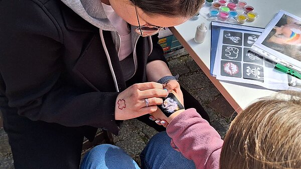 Eine Frau klebt ein Tattoo auf die Hand eines Kindes. Auf dem Tisch liegen Schablonen und Farben für das Schminken.