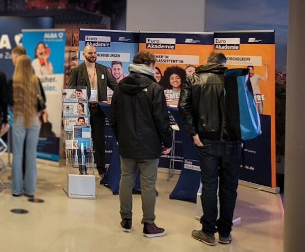 Rückblick auf die Jobmesse Leipzig Mehrere Personen stehen vor dem Messestand der Euro Akademie auf einer Jobmesse und sprechen mit einem Vertreter der Akademie, der einen Anzug trägt. Im Hintergrund sind drei große, bunte Banner mit Informationen zu verschiedenen Ausbildungs- und Studienmöglichkeiten zu sehen. Ein Prospektständer mit Infomaterial steht links neben dem Stand. Die Besucher tragen Jacken und Rucksäcke, während sie sich über Bildungsangebote informieren.