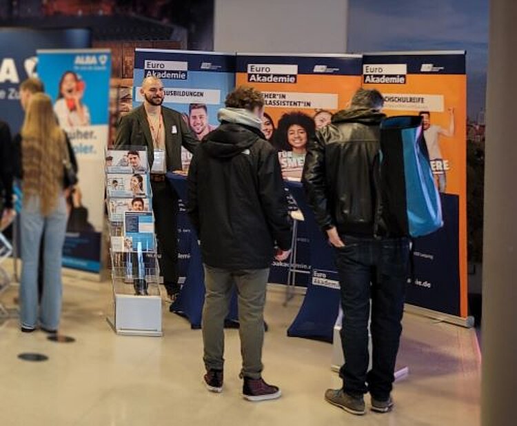 Rückblick auf die Jobmesse Leipzig Mehrere Personen stehen vor dem Messestand der Euro Akademie auf einer Jobmesse und sprechen mit einem Vertreter der Akademie, der einen Anzug trägt. Im Hintergrund sind drei große, bunte Banner mit Informationen zu verschiedenen Ausbildungs- und Studienmöglichkeiten zu sehen. Ein Prospektständer mit Infomaterial steht links neben dem Stand. Die Besucher tragen Jacken und Rucksäcke, während sie sich über Bildungsangebote informieren.