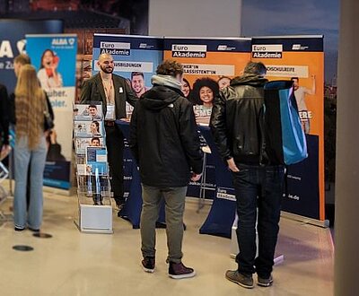 Mehrere Personen stehen vor dem Messestand der Euro Akademie auf einer Jobmesse und sprechen mit einem Vertreter der Akademie, der einen Anzug trägt. Im Hintergrund sind drei große, bunte Banner mit Informationen zu verschiedenen Ausbildungs- und Studienmöglichkeiten zu sehen. Ein Prospektständer mit Infomaterial steht links neben dem Stand. Die Besucher tragen Jacken und Rucksäcke, während sie sich über Bildungsangebote informieren.