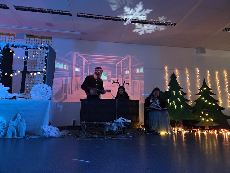 Bühnenbild mit winterlicher Scheunen-Projektion, links ein verschneiter Tisch, in der Mitte eine Person mit Rentiergeweih-Requisit an einem Pult, rechts zwei Personen vor beleuchteten Tannenbäumen und Lichterketten.