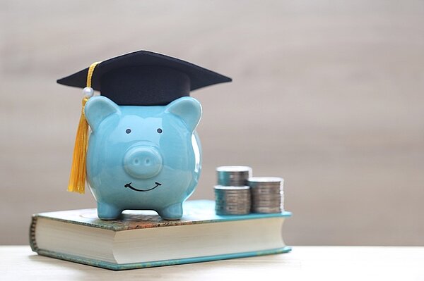 Bewirb dich für unser Stipendium an der Euro Akademie Nürnberg! Ein hellblaues, freundlich lächelndes Sparschwein steht auf einem Buch, neben ihm drei Türme von Münzen und auf seinem Kopf ein Graduation-Hut mit Zipfel.
