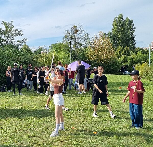 Wikinger-Sportfest: Pflegeazubis erobern die nordische Arena Junge Menschen stehen auf einer Wiese. Im Vordergrund spannt eine Person mit Brille und weißer Hose einen Bogen, während andere zuschauen und lachen.