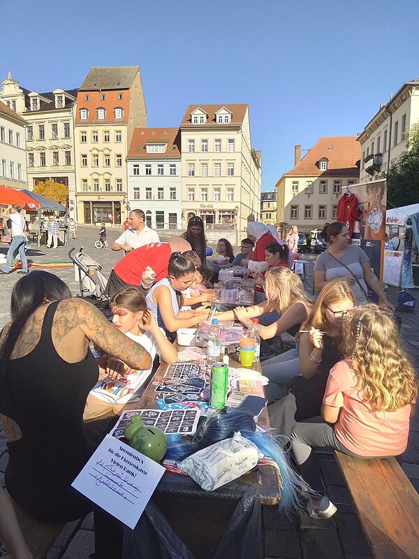 Nahaufnahme eines Basteltisches mit vielen Kindern und Erwachsenen, die malen, kleben und schminken, im Vordergrund bunte Materialien.