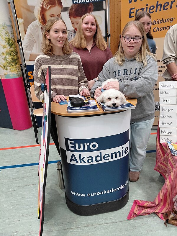 Auf dem Bild stehen die drei Auszubildenden Euro Akademie Tauberbischofsheim gemeinsam hinterm Stehpult des Euro-Akademie-Messestandes auf der Ausbildungsmesse, ein kleines weißes Hündchen auf dem Pult sitzend, und posieren für ein Erinnerungsbild.