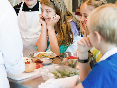 Auf diesem Bild bereiten Kinder und Erwachsene gemeinsam Essen zu. Eine schöne Szene, die das Lernen und die Verbindung durch kulinarische Künste hervorhebt.