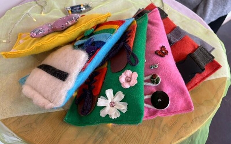Eine Sammlung von bunten, selbstgemachten Stofftaschen mit verschiedenen Mustern und Applikationen, darunter Blumen, Schmetterlinge und geometrische Formen, die auf einem Tisch liegen.