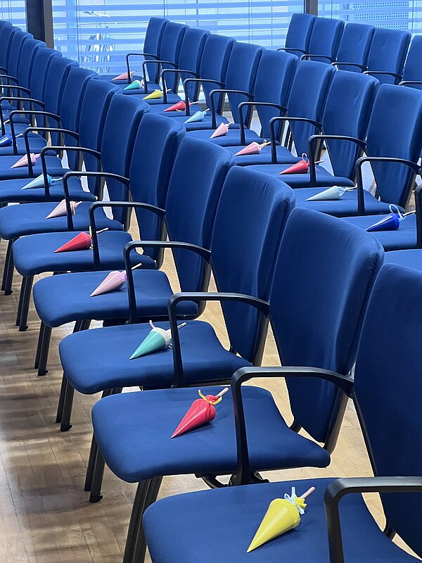 Reihen blauer Stühle in einem Veranstaltungssaal, auf denen kleine bunte Schultüten als Dekoration oder Geschenk bereitliegen.