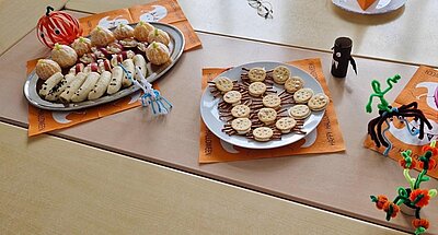 Halloween-Snackteller Auf einem Tisch, auf dem orangefarbene Halloween-Servietten und etwas Dekoration liegt, sind große Teller platziert, auf denen Salzstangen und Kräcker wie kleine Spinnengeister aussehen, Mandarinen wie kleine Kürbisse angerichtet sind, aus Apfelscheiben Vampirgebisse mit Zähnen und aus Bananen kleine Geister liegen.