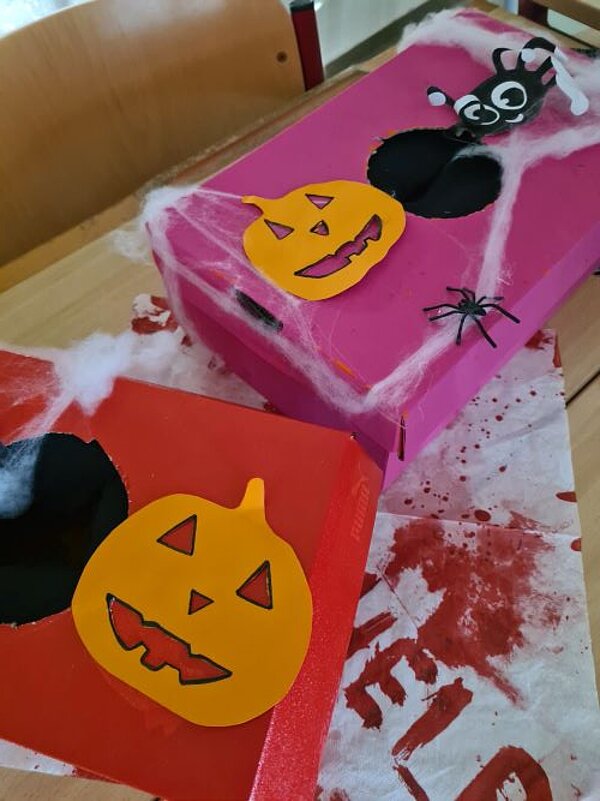 Zwei Schuhkartons, die mit einer runden Öffnung in der Mitte versehen sind und mit Halloween-Kürbissen aus Papier, Dekospinnen und Spinnweben aus Watte dekoriert sind.