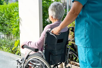 Eine Pflegekraft in blauer Arbeitskleidung schiebt eine ältere Person im Rollstuhl nach draußen. Die Szene spielt in einem grünen, gepflegten Außenbereich, was auf eine unterstützende und fürsorgliche Umgebung hinweist.