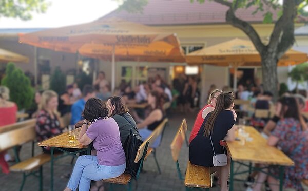 Wiedersehen und Erinnerungen: Ein unvergessliches Alumnitreffen Absolventinnen und Absolventen stehen und sitzen bei sommerlichem Wetter draußen auf Bänken an Tischen unter Sonnenschirmen.