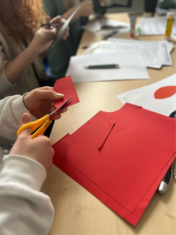 Mathe im Kindergarten? Geht und macht Spaß! Die Schüler*innen schneiden mit Scheren aus bunten Pappen verschiedene Formen aus. Auf dem Tisch stehen außerdem Klebestifte.