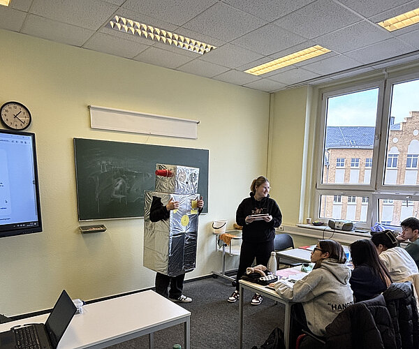 Ein Schüler in Roboterverkleidung steht vorne im Klassenzimmer neben einer Mitschülerin. Mehrere Schüler sitzen an ihren Plätzen und schauen interessiert zu. Rechts sieht man ein großes Fenster mit Blick auf ein Schulgebäude.