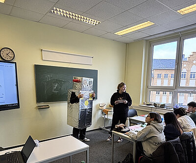 Ein Schüler in Roboterverkleidung steht vorne im Klassenzimmer neben einer Mitschülerin. Mehrere Schüler sitzen an ihren Plätzen und schauen interessiert zu. Rechts sieht man ein großes Fenster mit Blick auf ein Schulgebäude.