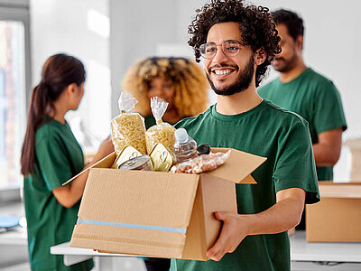 Ein junger Mann mit Brille und grünem T-Shirt trägt einen Karton mit Lebensmitteln und lächelt. Andere Personen mit grünen T-Shirts stehen im Hintergrund und scheinen Kartons zu packen.