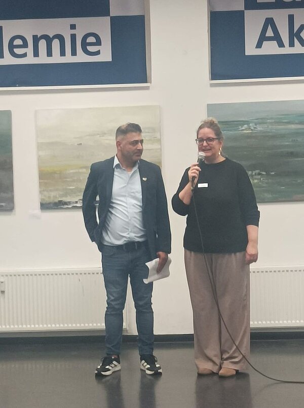 Eine Frau und ein Mann stehen vor einer Wand, an der abstrakte Gemälde und Schilder mit "Akademie" hängen. Die Frau spricht in ein Mikrophon.