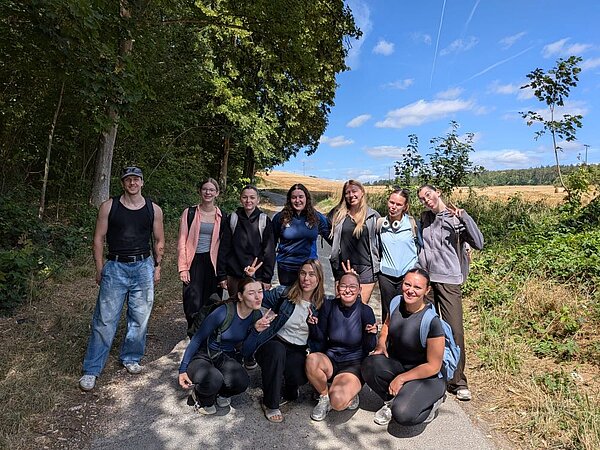 Eine Gruppe von elf Personen posiert auf einem Waldweg. Die meisten lächeln in die Kamera, während sie in einer lockeren Formation stehen oder knien. Einige machen das Peacezeichen und strecken die Zunge heraus. Im Hintergrund sind Bäume und Felder zu sehen.