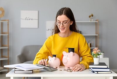 Junge Frau am Schreibtisch zählt lächelnd Geld, vor ihr stehen zwei Sparschweine, Bücher und ein Taschenrechner.