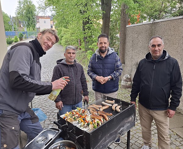 Vier Männer stehen im Freien an einem Grill mit Würstchen und Gemüsespießen.