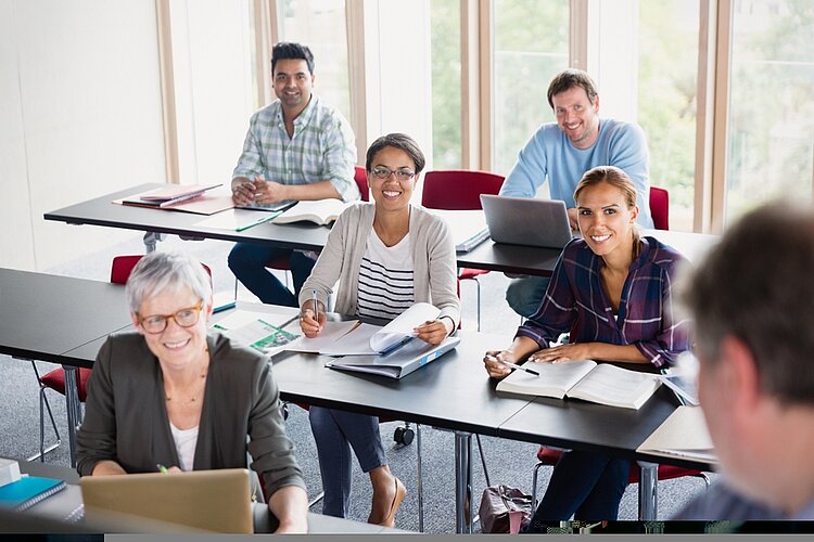 Fünf Personen sitzen an aufgereihten Tischen und hören einem Sprecher vorne zu. Ganz ganz hinten sitzen zwei Männer, in der Mitte zwei junge Frauen und ganz vorne eine ältere Frau.
