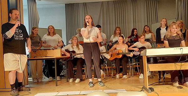 Schulband steht auf der Bühne und performt für das Publikum. Eine Sängerin, ein Sänger, eine Keyboarderin, zwei am Xylophon und mehrere an Akustikgitarren begleiten den Gesang instrumental.