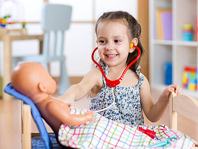Ein kleines Mädchen mit einem Stethoskop um den Hals untersucht eine Puppe, die auf einem kleinen Bett liegt. Das Mädchen lächelt und scheint in einem Spielzimmer zu sein, das mit verschiedenen Spielsachen ausgestattet ist.