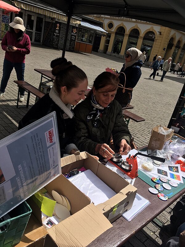 Protesttag zur Gleichstellung von Menschen mit Behinderung: Buttons für Inklusion Zwei Frauen unterschiedlichen Alters sitzen an einem Tisch, auf dem eine Buttonmaschine steht. Auf dem Tisch befinden sich mehrere Utensilien. Im Hintergrund sieht man Passante*innen über den Würzburger Marktplatz laufen.