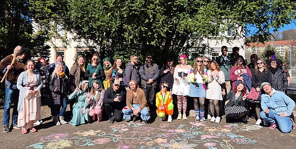 Eine große Gruppe Schülerinnen steht draußen vor einem Gebäude und posiert in verschiedenen bunten Kostümen passend zum Motto „Zeitreise“. Auf dem Boden ist bunte Kreidekunst mit „ESO“ zu sehen.*