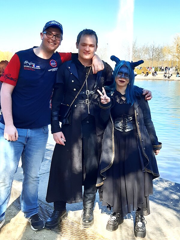 Buchmesse Leipzig: Wo Worte Bildung und Austausch fördern Drei Personen posieren fröhlich vor einer Wasserfontäne, umgeben von vielen Menschen. Zwei von ihnen tragen auffällige, düstere Outfits mit Fantasy- oder Gothic-Elementen, während die dritte Person sportlich-leger gekleidet ist. Die Szene ist auf der Leipziger Buchmesse aufgenommen.