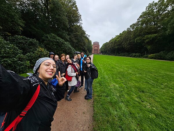 Klassenausflug Planetarium KASS23 Auf einem Weg in einem Park steht eine Schülergruppe. Ganz vorne links macht die junge Frau, die das Selfie auslöst, ein Peace-Zeichen. Hinten im Bild ist ein altes Gebäude mit einer Kuppel erkennbar.