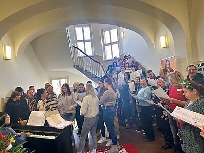 Eine Gruppe ist in einem Treppenhaus versammelt und singt gemeinsam Weihnachtslieder. Die Personen halten Notenblätter in den Händen. Auf der linken Seite spielt eine Frau an einem Klavier. Es ist ein kleines Stück von einem Weihnachtsbaum zu sehen.